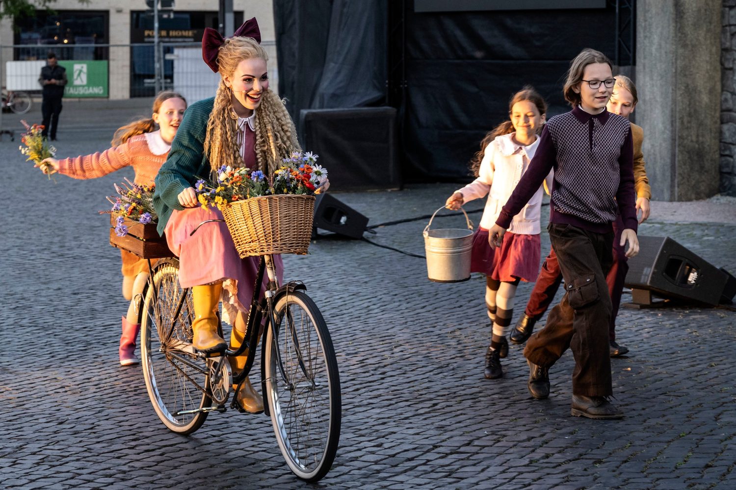 Alexandra Yangel riding a bicycle with a basket of wildflowers, children running behind her