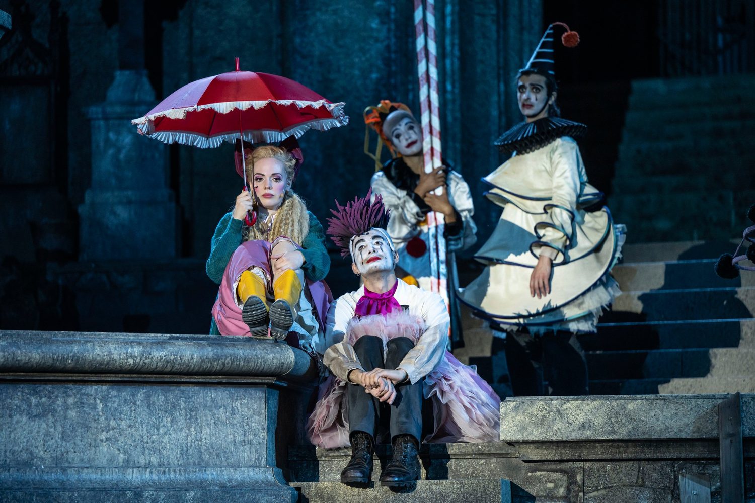 Alexandra Yangel under a red parasol beside performers in clown make-up