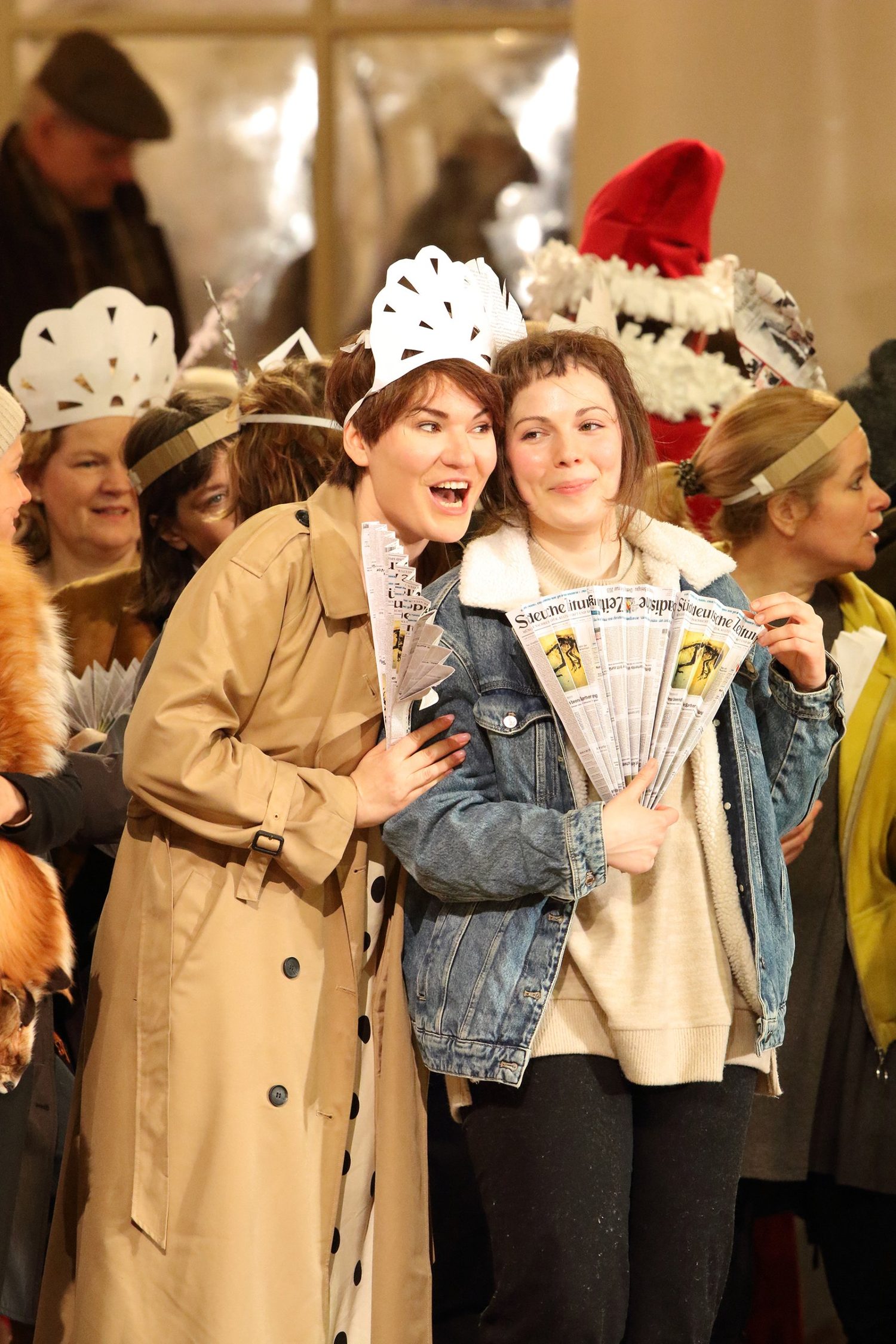 Alexandra Yangel holding a fan of rolled newspapers, cheek to cheek with another singer in a paper crown